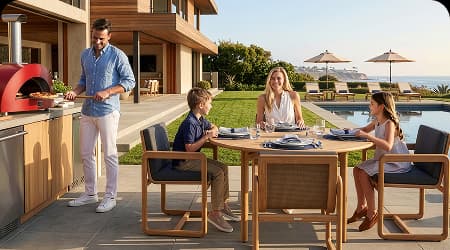 Family dining at a teak outdoor table beside a pool while a man prepares pizza in a red outdoor oven, with ocean views beyond the patio.