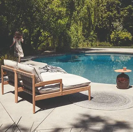 A stylish outdoor lounge set featuring a wooden loveseat and armchairs with woven cord backs and light grey cushions is arranged on a modern stone patio next to a swimming pool.