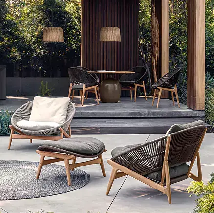 A stylish outdoor lounge area features a woven loveseat, matching rounded lounge chairs, and an ottoman with natural wood frames arranged on a concrete patio with round woven rugs and lush garden landscaping.