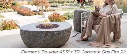 Elementi Boulder concrete gas fire pit with lava rock beside a woman reading on a modern patio.